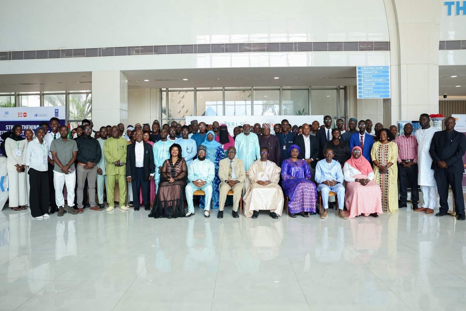 A large group of people posed together for a photograph in a conference centre lobby, with an SRLS banner and event signage from partner organisations behind them.