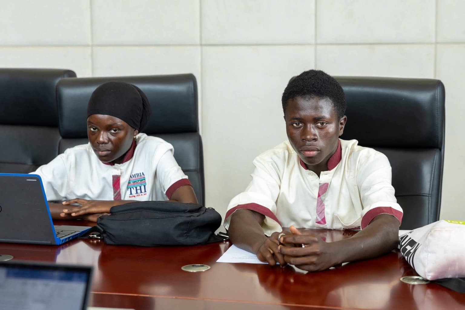 Two young people sitting at a conference table with a laptop and papers, wearing matching branded shirts, in an office meeting room.