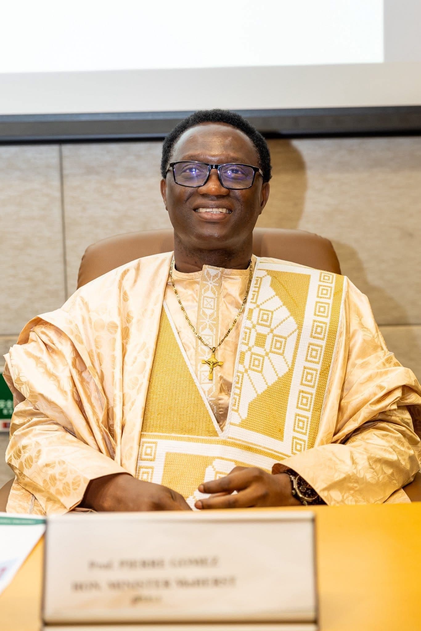 A man in ceremonial Gambian dress sitting at a desk with a nameplate in front of him.