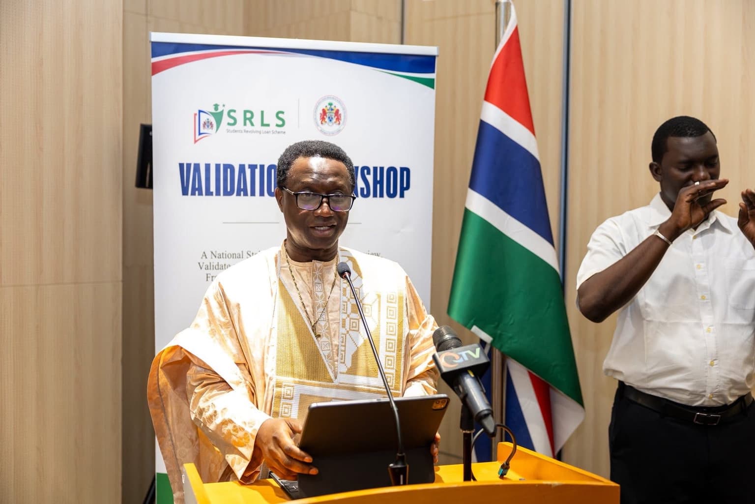 A man speaking at a podium in front of an SRLS banner reading Validation Workshop, beside the Gambian flag, with another man standing nearby.