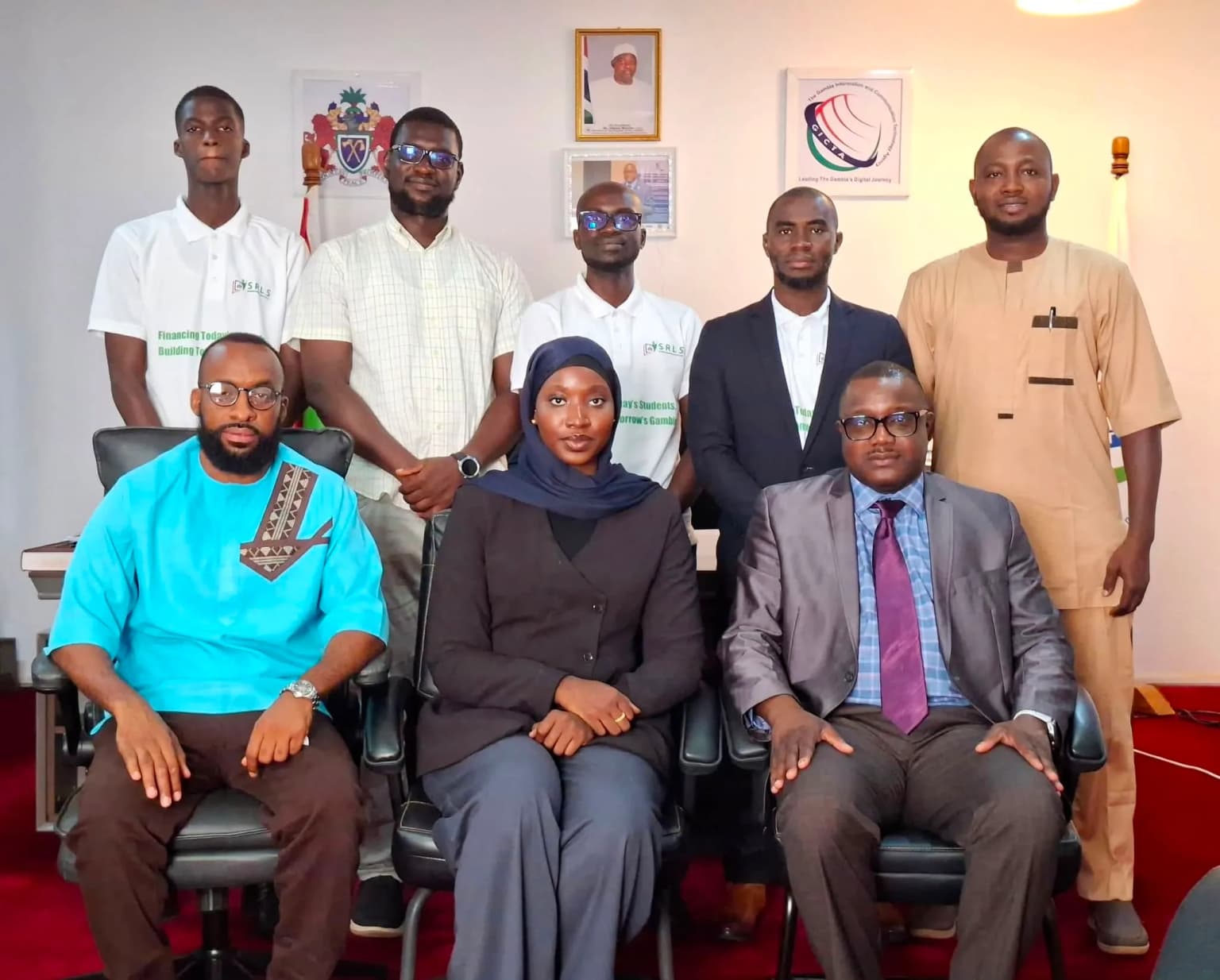 Eight members of the SRLS team photographed together at a government office, under the coat of arms of The Gambia.